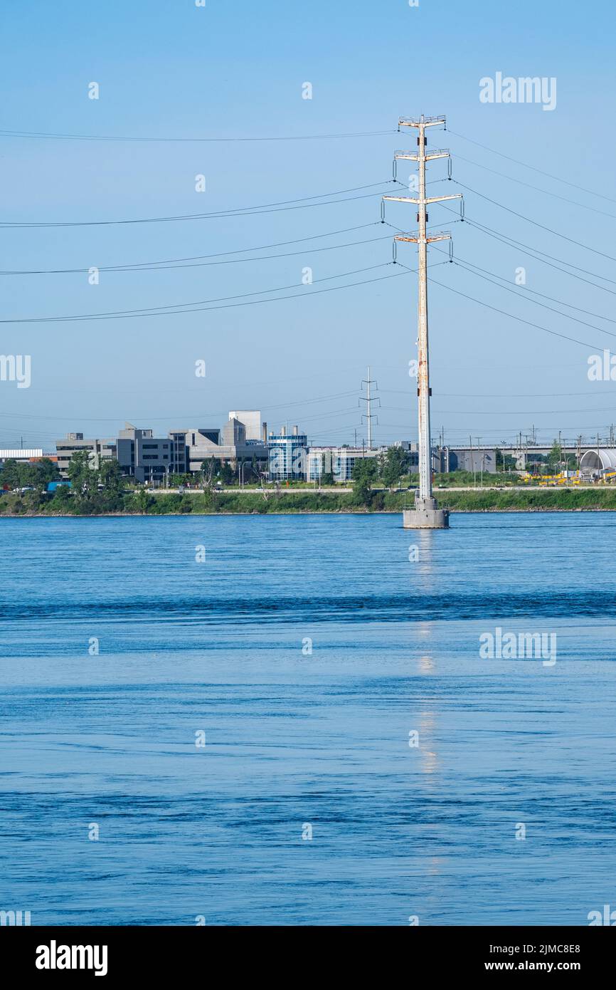 An electric power distribution pole stands in a river with buildings on a shore in the distance. Stock Photo