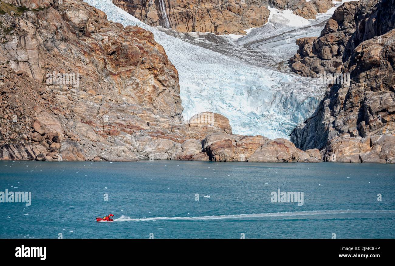 Small red boat crossing in front of massive front of a glacier in Prince Christian Sound, South Greenland on 22 July 2022 Stock Photo
