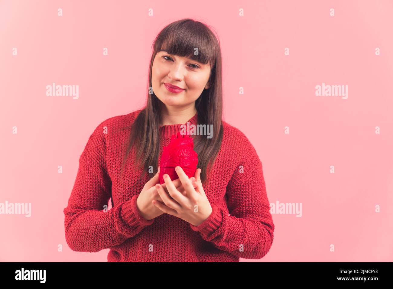 Natural-looking relaxed mature woman with long straight brown hair, holding an artificial red heart in front of her chest, reminding about health problems, illness, and cardiovascular diseases. Medium studio shot with a pink background. High quality photo Stock Photo