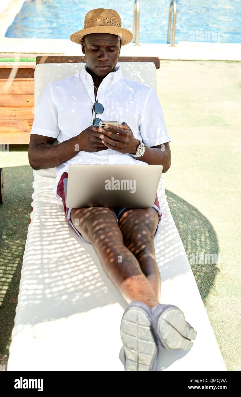 Full body of African American businessman with netbook surfing cellphone while lying on lounger under parasol in coastal area on summer day Stock Photo
