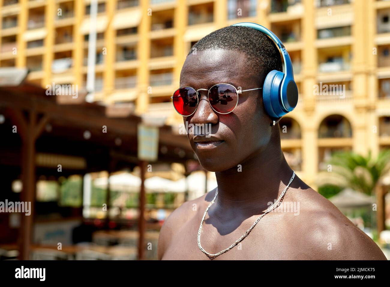 Confident African American shirtless male listening to song in wireless headphones while standing near swimming pool in tropical resort on summer day Stock Photo