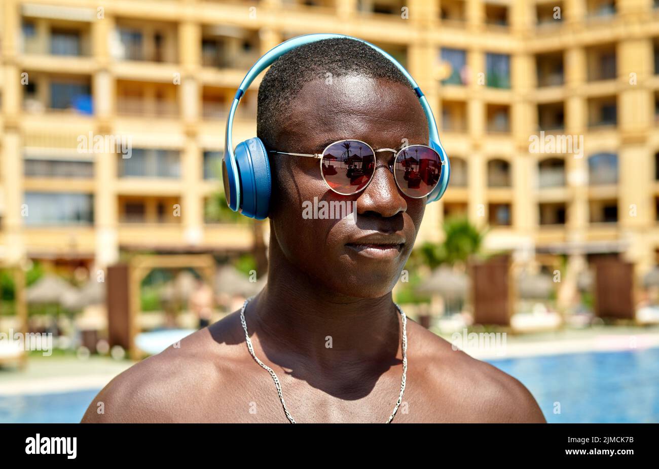 Confident African American shirtless male listening to song in wireless headphones while standing near swimming pool in tropical resort on summer day Stock Photo