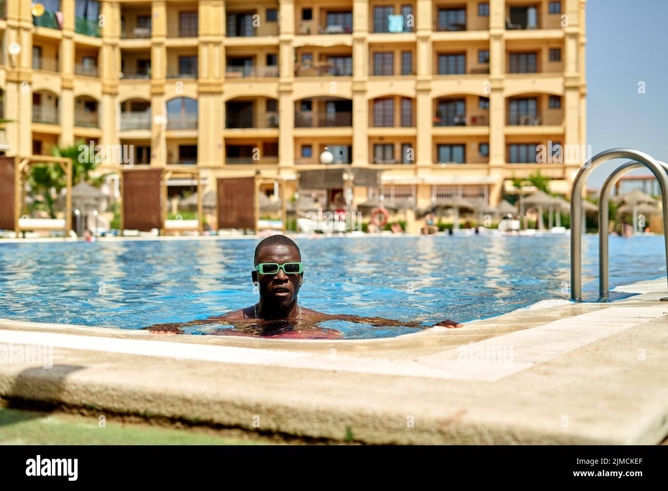 Confident shirtless African American male in sunglasses standing in swimming pool during summer vacation in tropical resort with multistory building Stock Photo