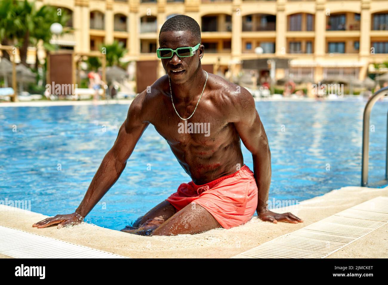 Confident shirtless African American male in sunglasses standing in swimming pool during summer vacation in tropical resort with multistory building Stock Photo