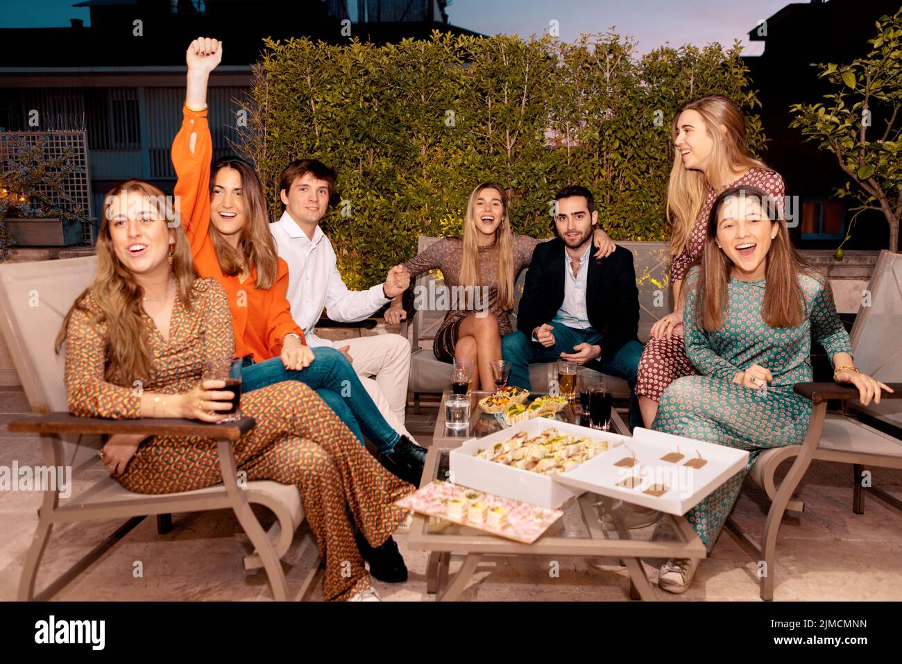 Group of positive friends celebrating festive event while gathering around table with appetizers during dinner in evening time on terrace Stock Photo