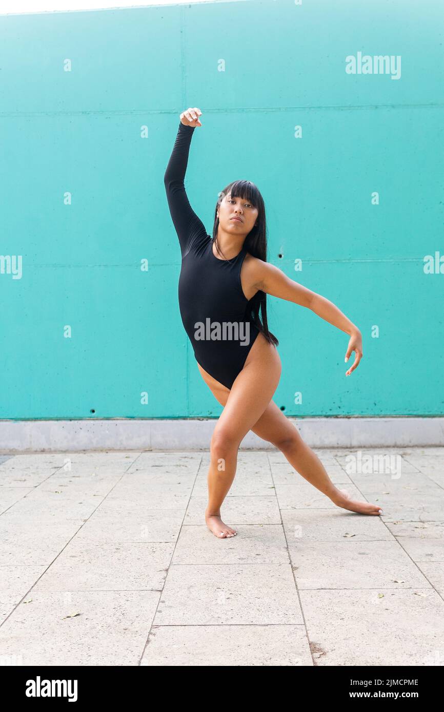 Full body barefoot ethnic woman in black leotard raising arms while dancing looking at camera on pavement against turquoise wall during performance on Stock Photo
