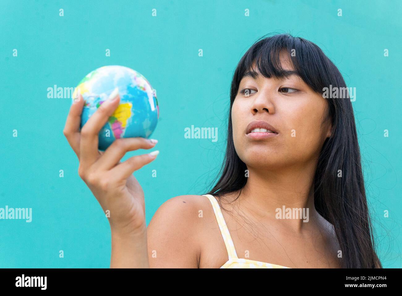 Thoughtful young ethnic female tourist with long hair examining small globe while planning trip against turquoise background Stock Photo