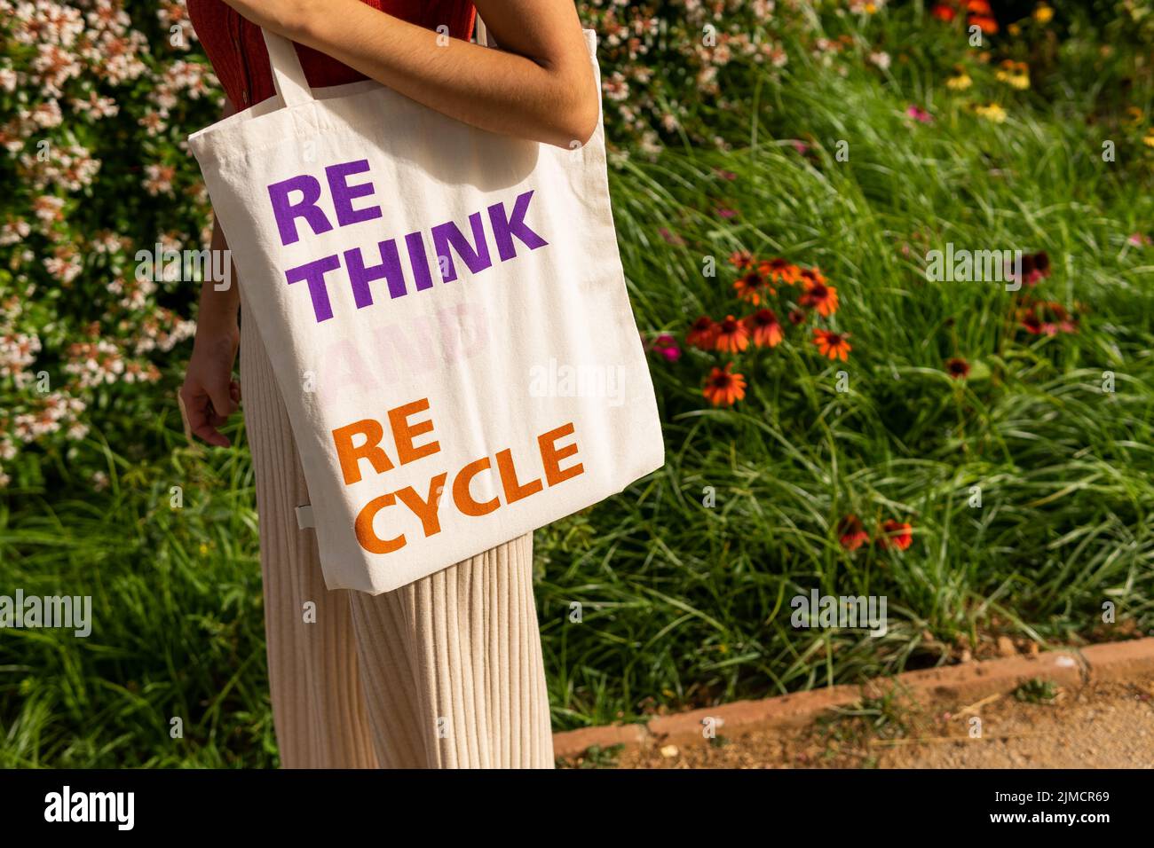 Anonymous female carrying ECO friendly fabric bag with Rethink Recycle inscription while standing near blooming bushes on street Stock Photo