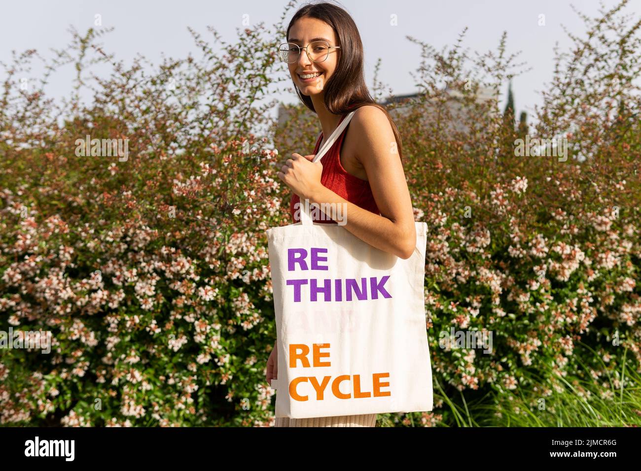 Positive female carrying ECO friendly fabric bag with Rethink Recycle inscription looking at camera while standing near blooming bushes on street Stock Photo