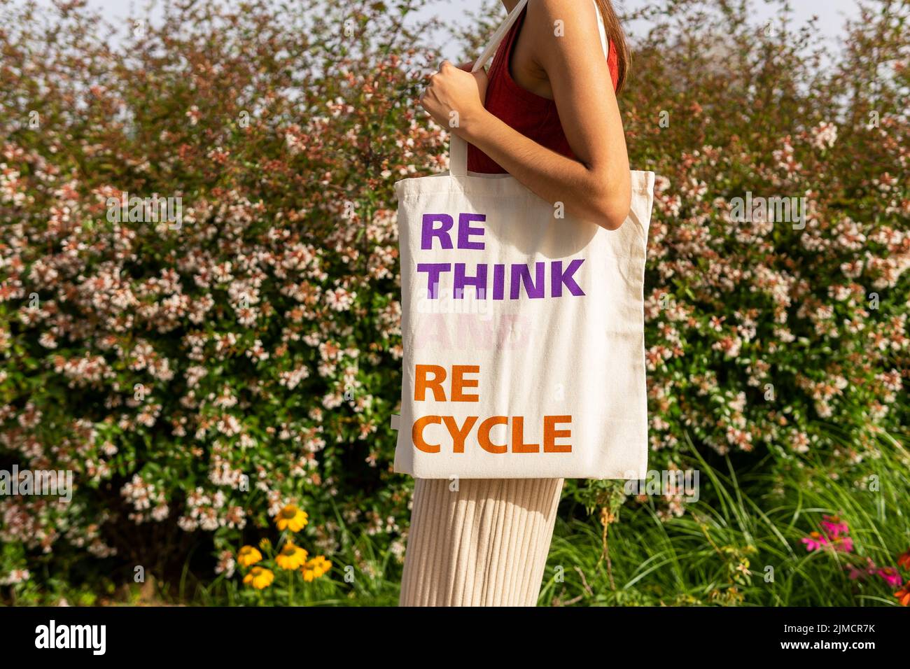 Anonymous female carrying ECO friendly fabric bag with Rethink Recycle inscription while standing near blooming bushes on street Stock Photo