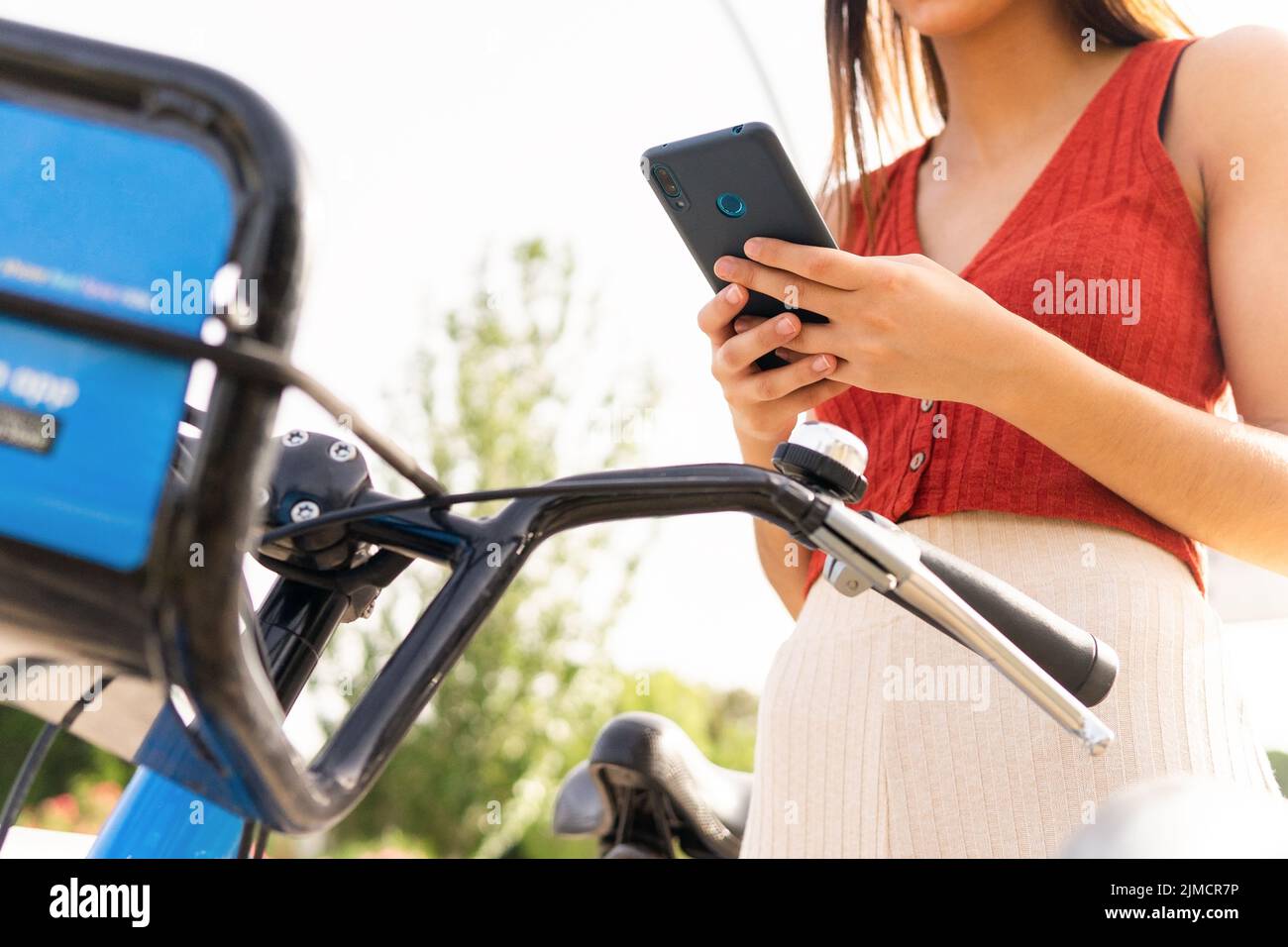 Anonymous female with backpack surfing modern cellphone while renting bicycle on street with buildings on sunny summer day in city Stock Photo