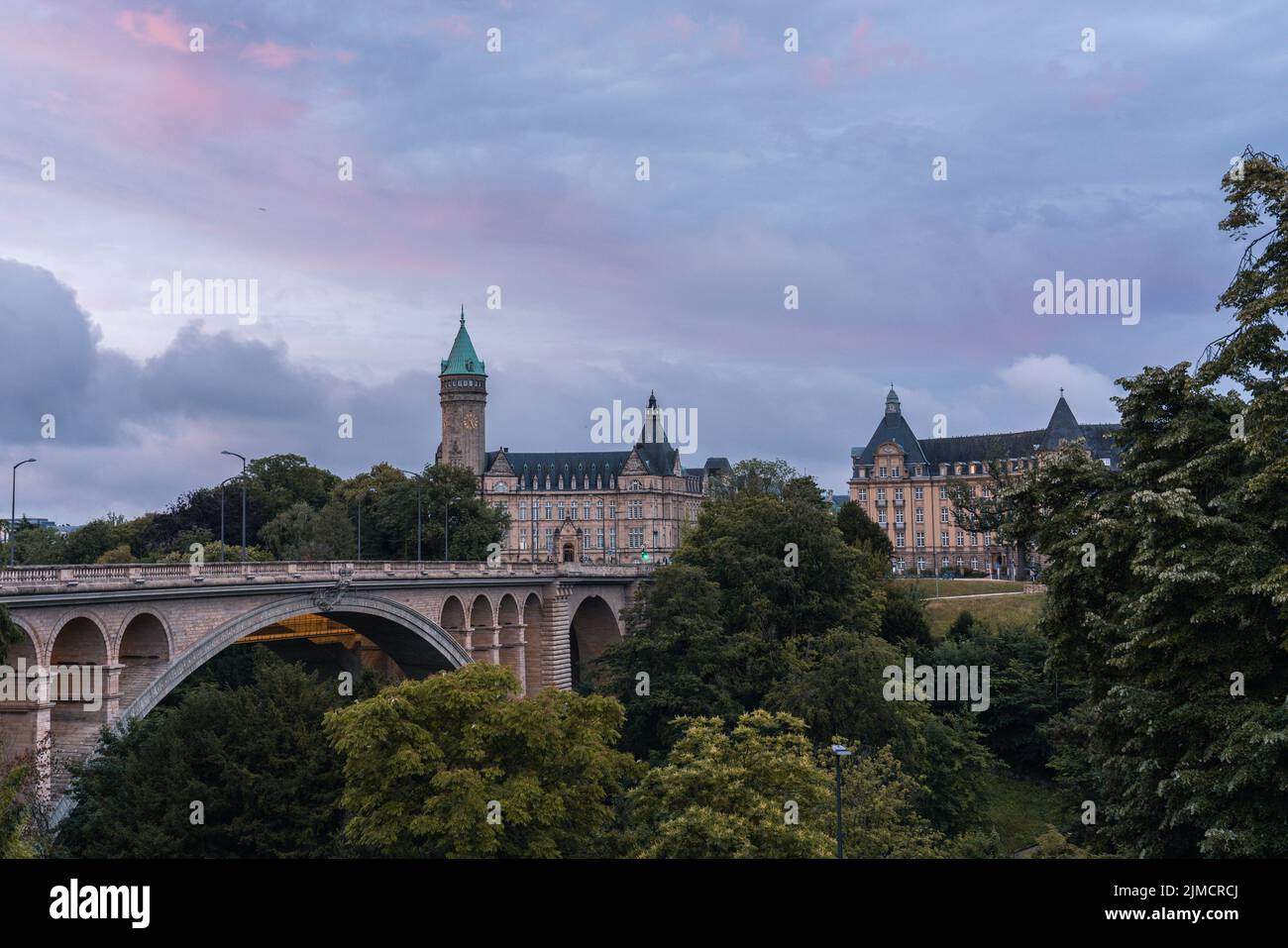Picturesque scenery of historic arched Adolphe Bridge surrounded by lush green trees leading to aged castle against cloudy sunset sky in Luxembourg Stock Photo