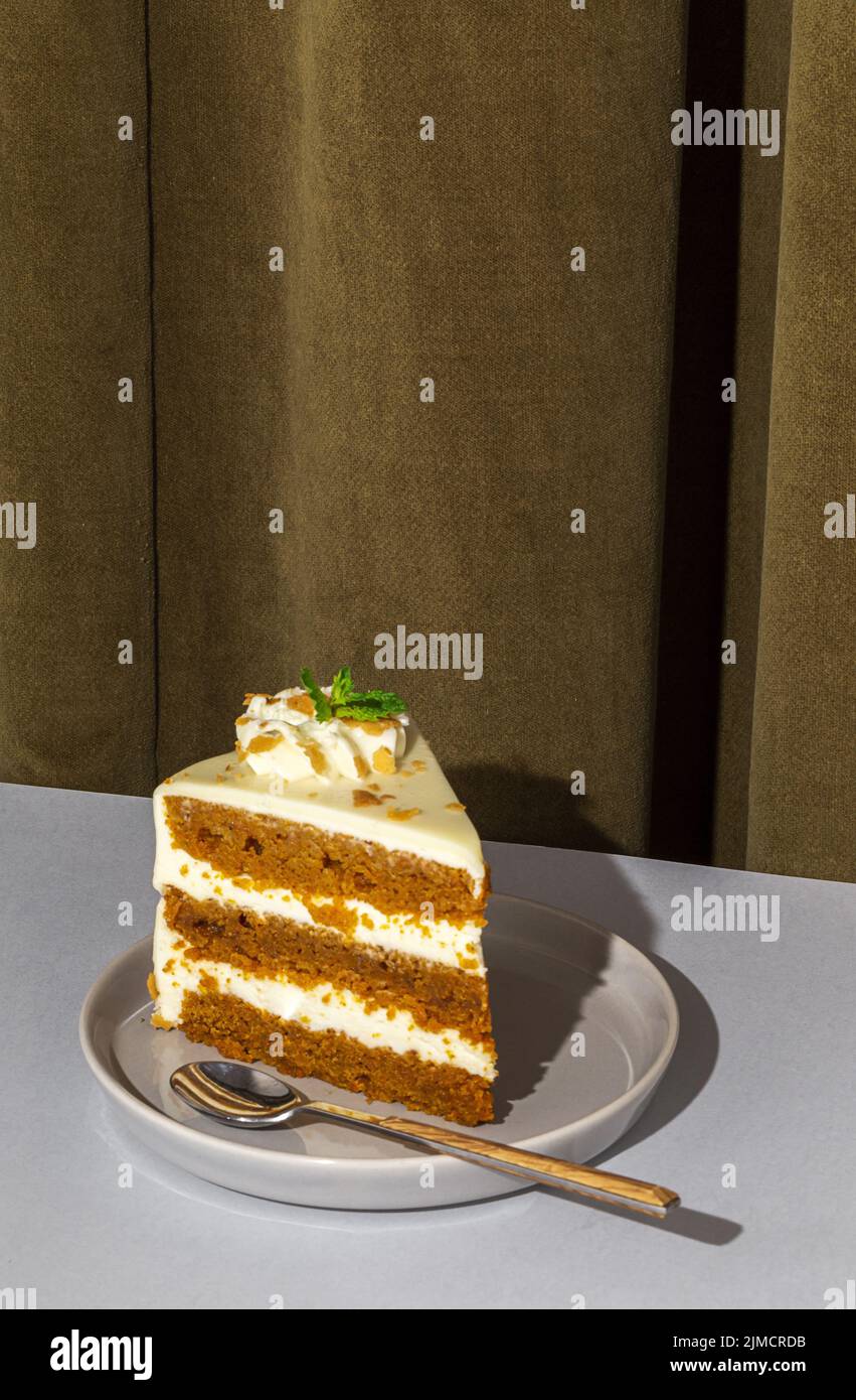 Slice of tasty sweet carrot sponge cake with cream decorated with mint leaf served on plate with spoon on table in light room with curtain on the back Stock Photo