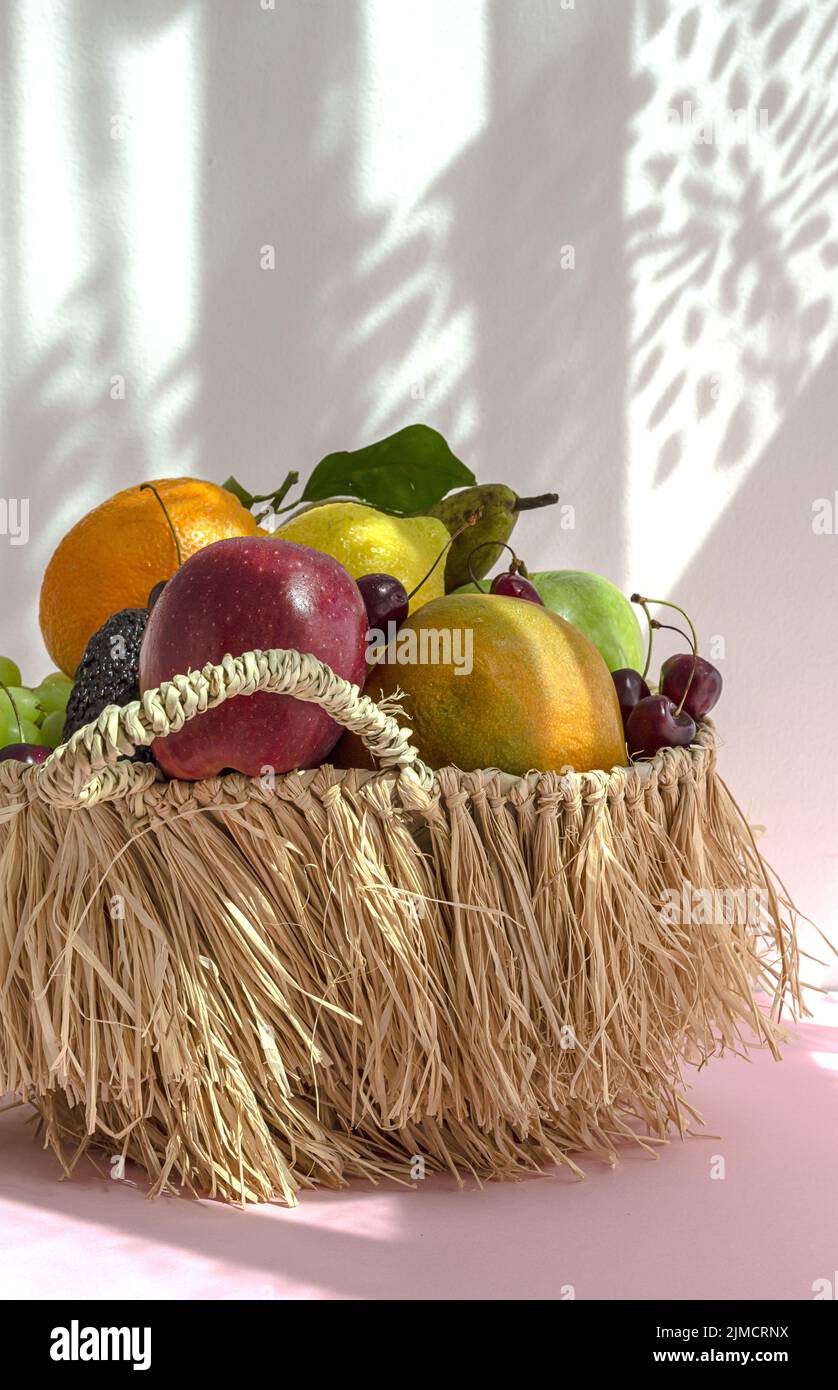 Still life of assorted ripe fruits and grapes placed in wicker basket with green mint leaves on table against white background with shadow Stock Photo