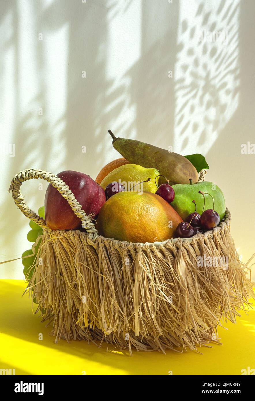Still life of assorted ripe fruits and grapes placed in wicker basket with green mint leaves on table against yellow and white background with shadow Stock Photo