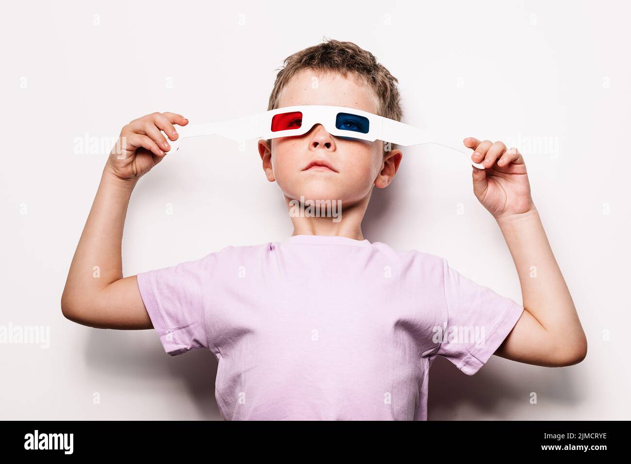 Cute boy in casual wearing 3D glasses with colorful lenses while standing looking away against white background in light studio Stock Photo