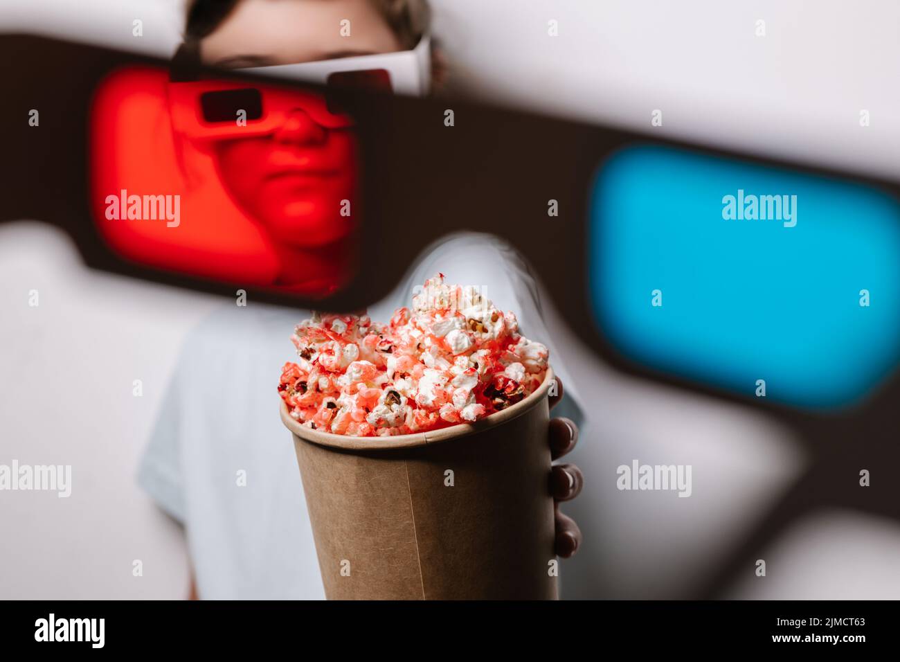 Through red lens of content girl in 3D glasses with tasty popcorn reaching hand to camera standing against gray background in light studio Stock Photo