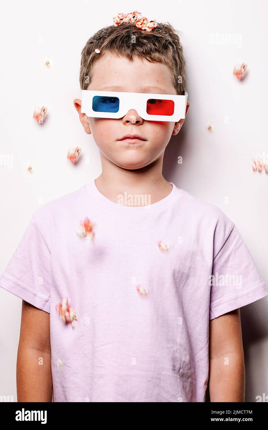Cute kid in 3D glasses with colorful lenses throwing popcorn over head while looking at camera standing against white background in light studio Stock Photo