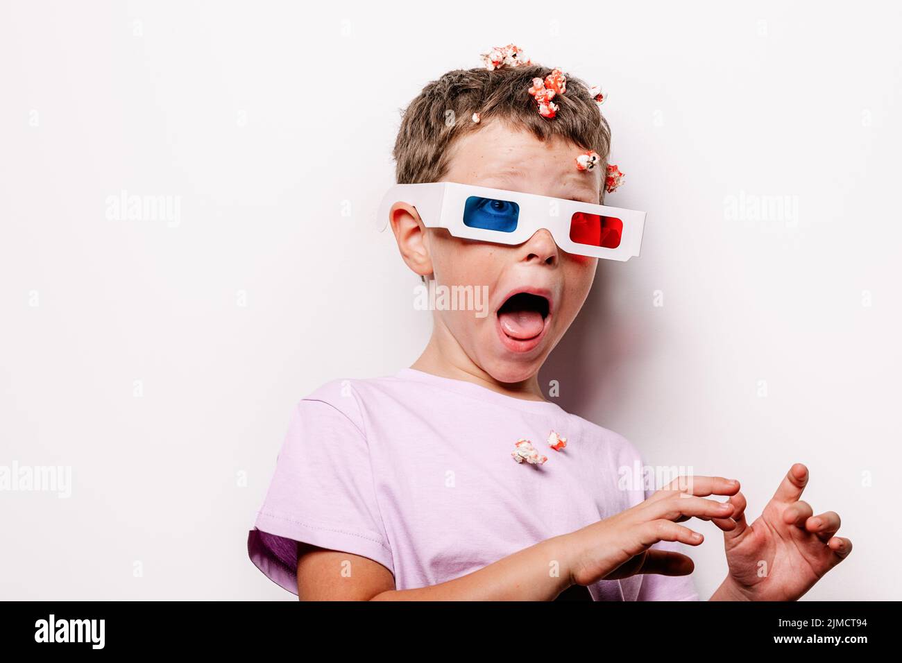 Child in 3D glasses with colorful lenses throwing tasty popcorn with opened mouth while standing against white background in light studio Stock Photo