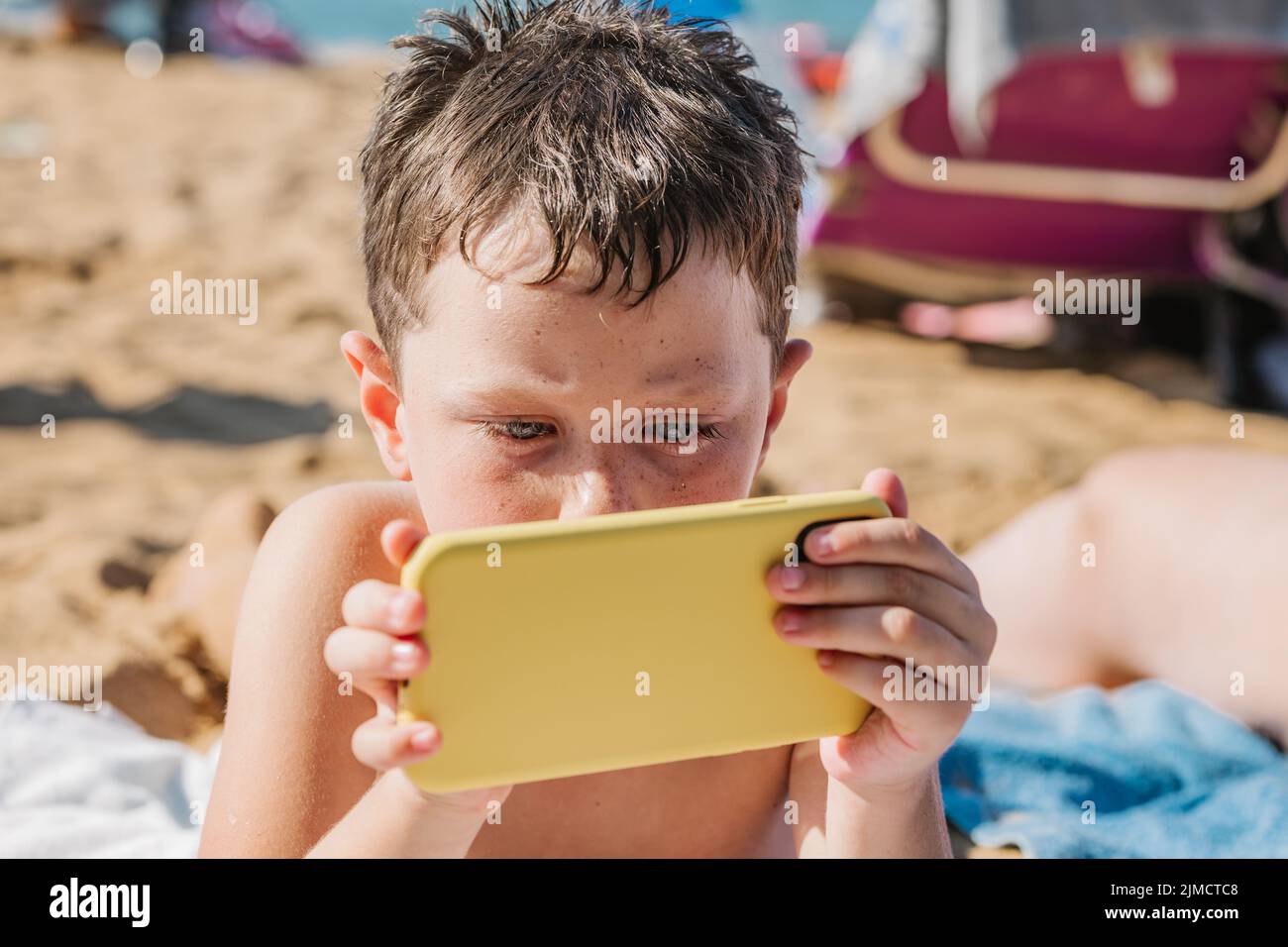 Focused boy in swimming shorts playing video game on cellphone while lying on sandy shore in tropical resort on sunny summer day in Getaria Zarautz Stock Photo