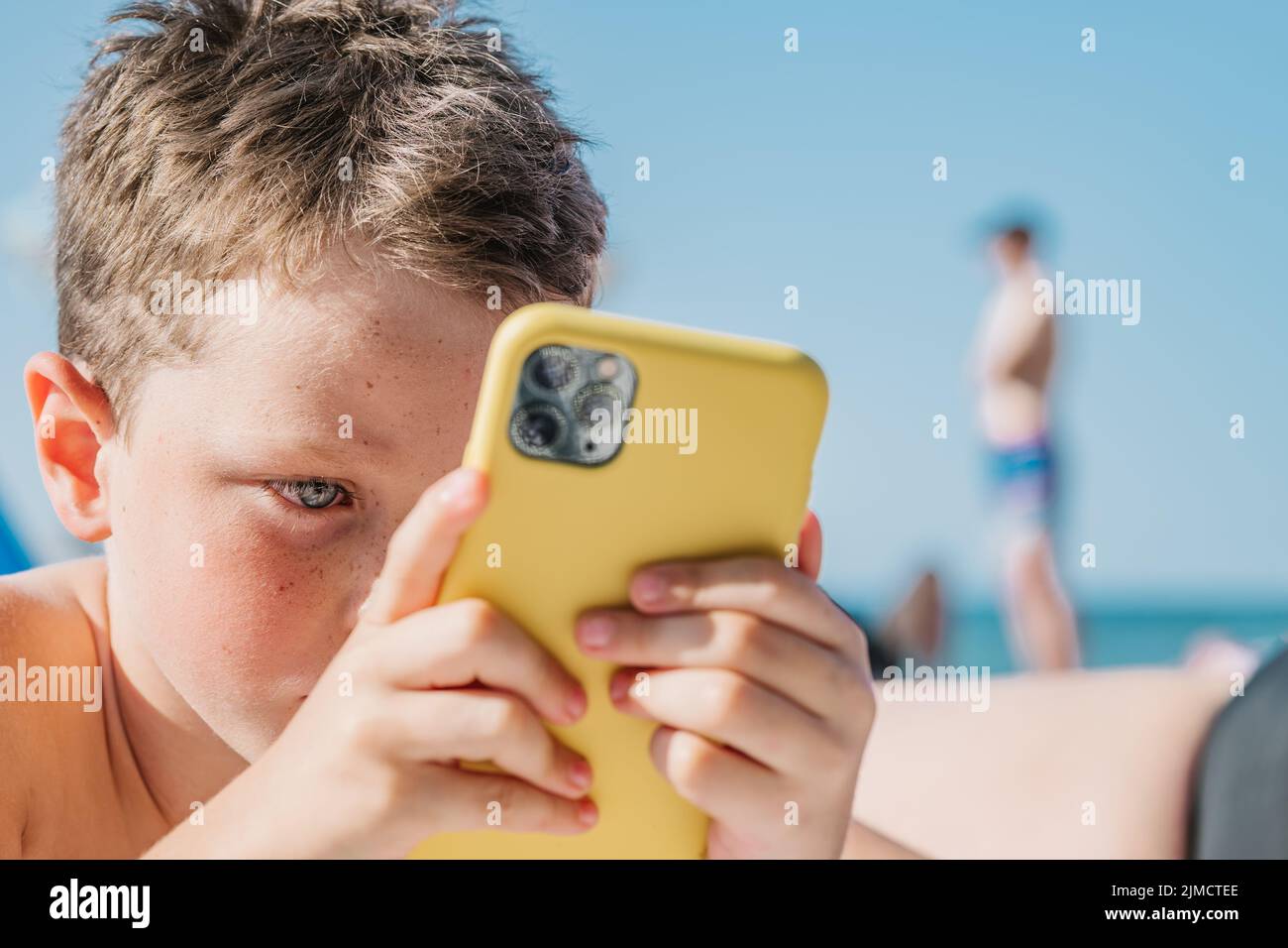 Focused boy in swimming shorts playing video game on cellphone while lying on sandy shore in tropical resort on sunny summer day in Getaria Zarautz Stock Photo