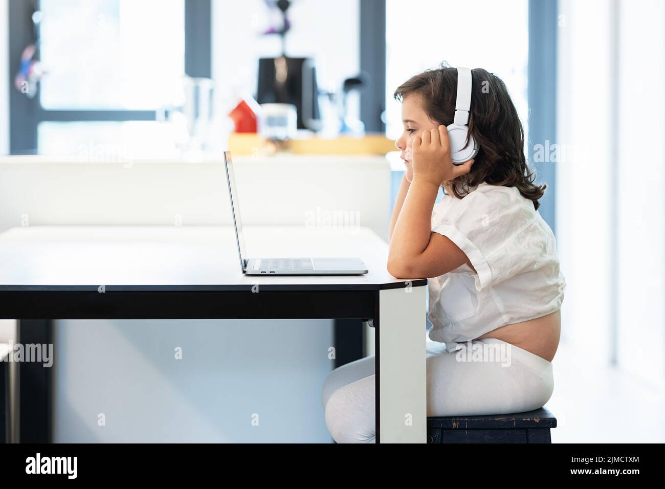 Side view of girl with a bored face and with headphones on her head sitting in front of a laptop doing home work looking at screen Stock Photo