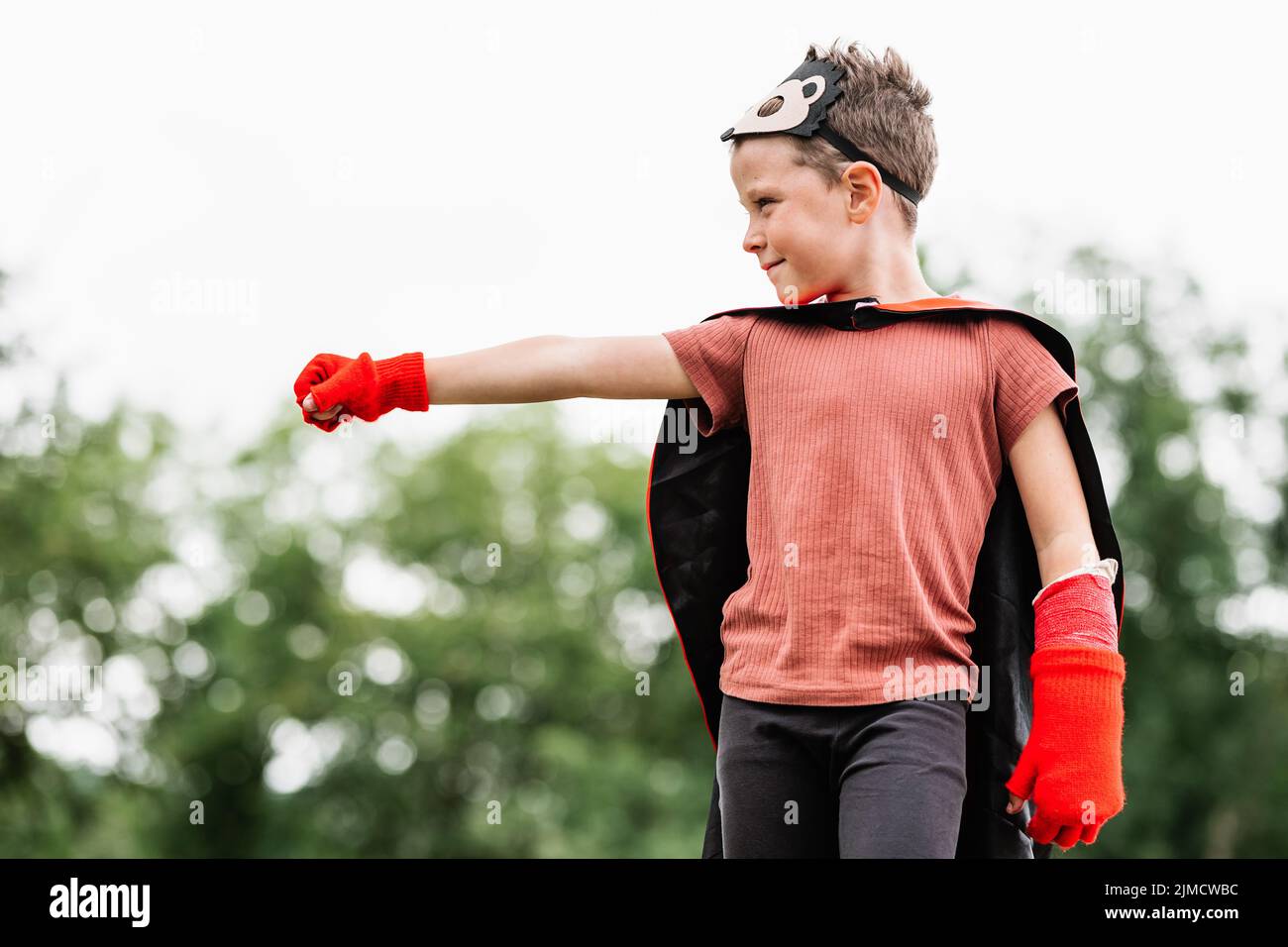 Side view of boy in red superhero cape and hedgehog mask looking away while standing with outstretch arm with clenched fist on stone block on blurred Stock Photo