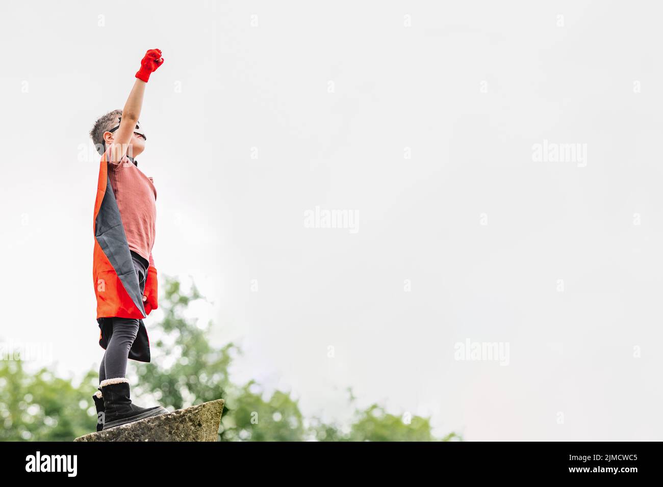 Side view of boy in red superhero cape and hedgehog mask looking away while standing with arms raised with clenched fist on stone block on blurred bac Stock Photo
