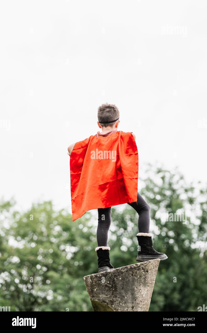 Back view of unrecognizable boy in red superhero cape and mask looking away while standing on stone block on blurred background of park trees Stock Photo