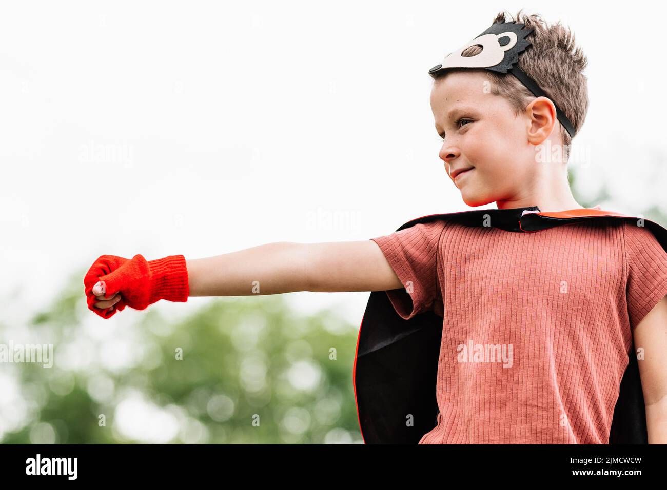 Side view of boy in red superhero cape and hedgehog mask looking away while standing with outstretch arm with clenched fist on stone block on blurred Stock Photo