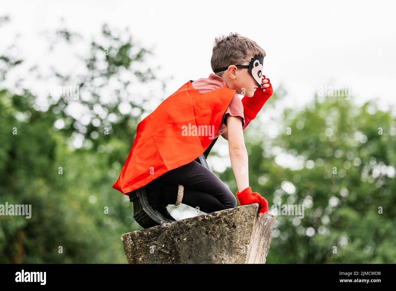 Side view of boy in red superhero cape and hedgehog mask touching face and looking away while kneeling on stone block on blurred background of park tr Stock Photo