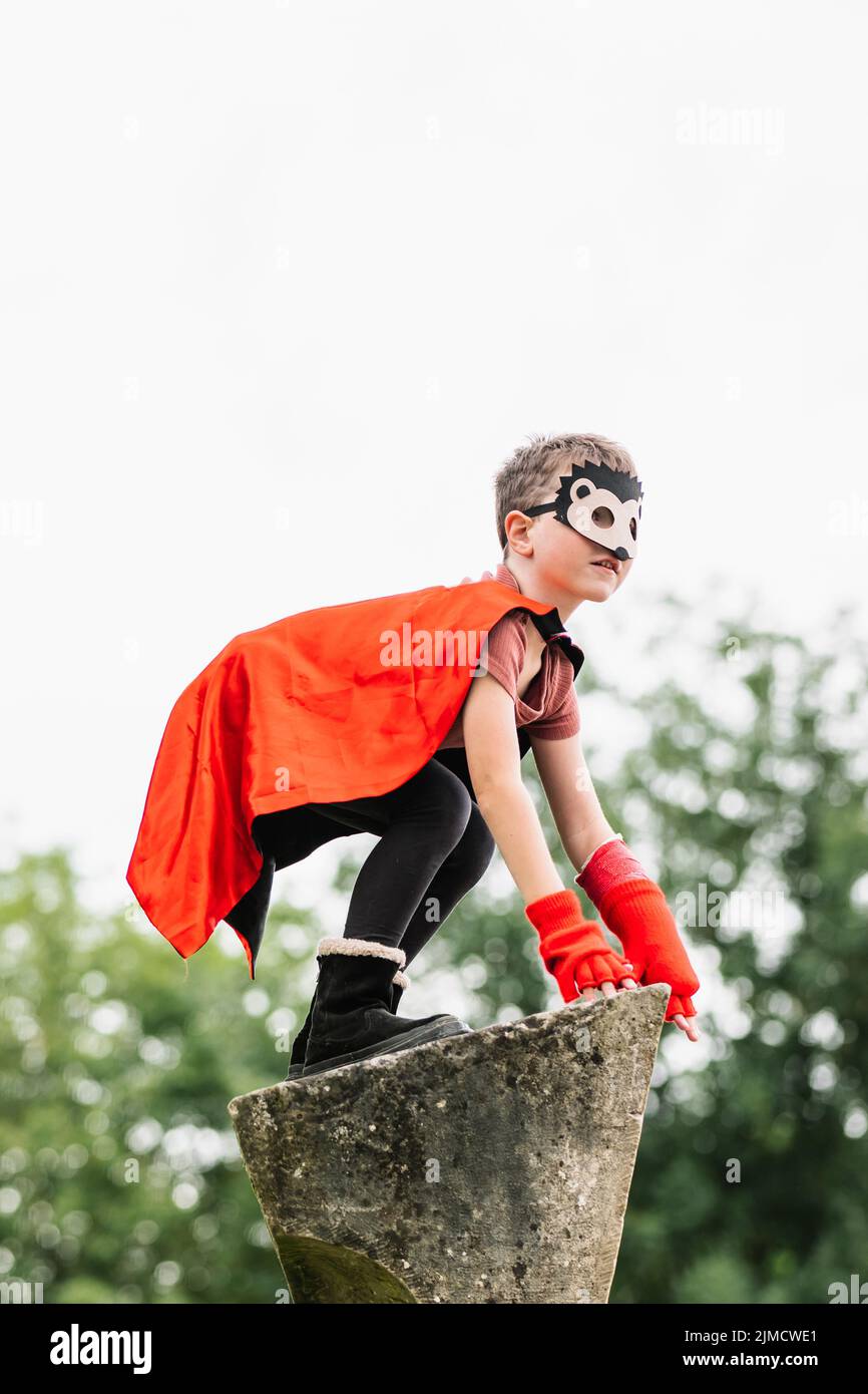 Side view of boy in red superhero cape and hedgehog mask looking away while standing on stone block on blurred background of park trees Stock Photo
