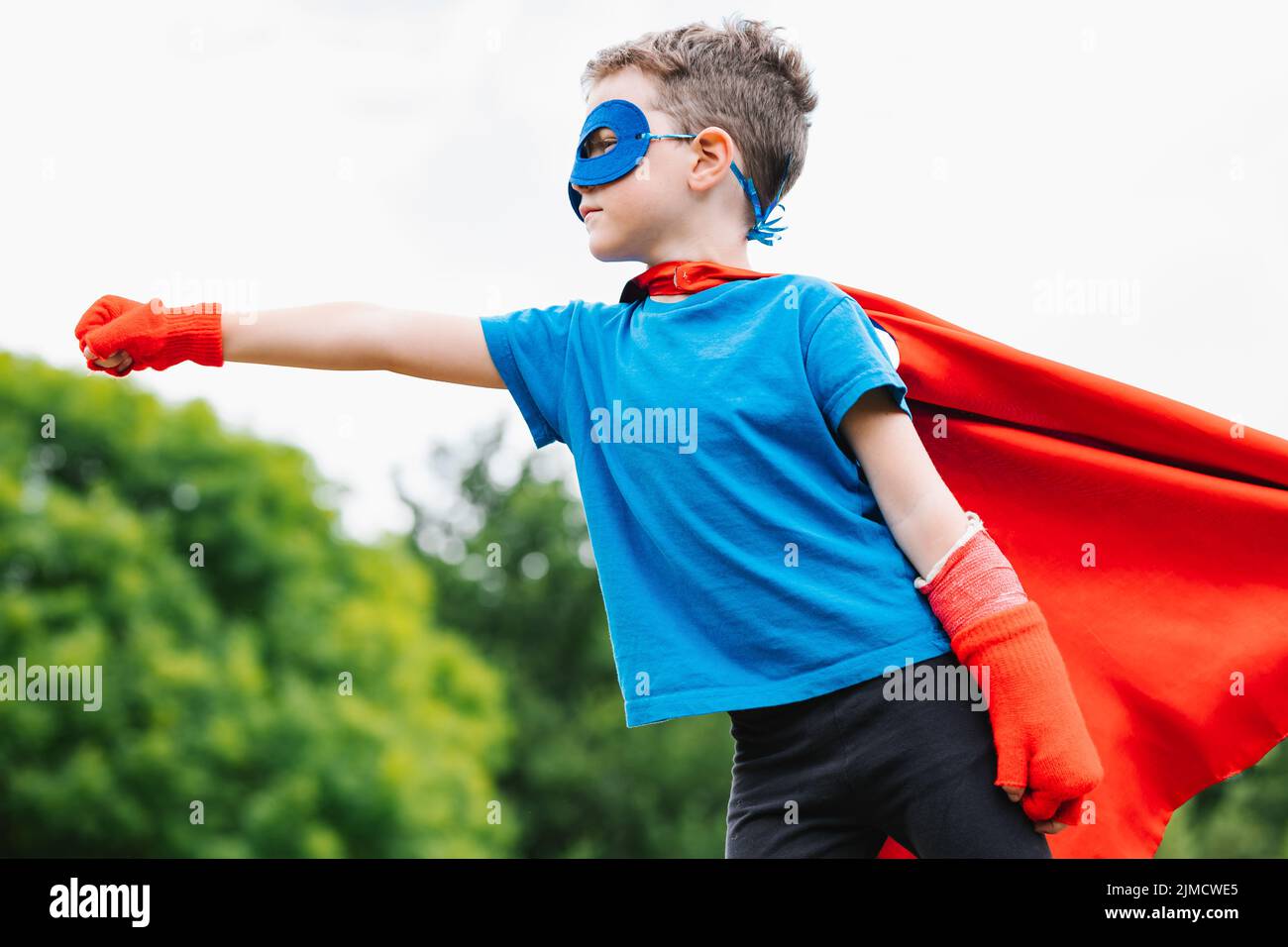 Side view of little superhero with cape and mask raising arm with clenched fist and screaming against green trees and cloudy sky on summer day in park Stock Photo