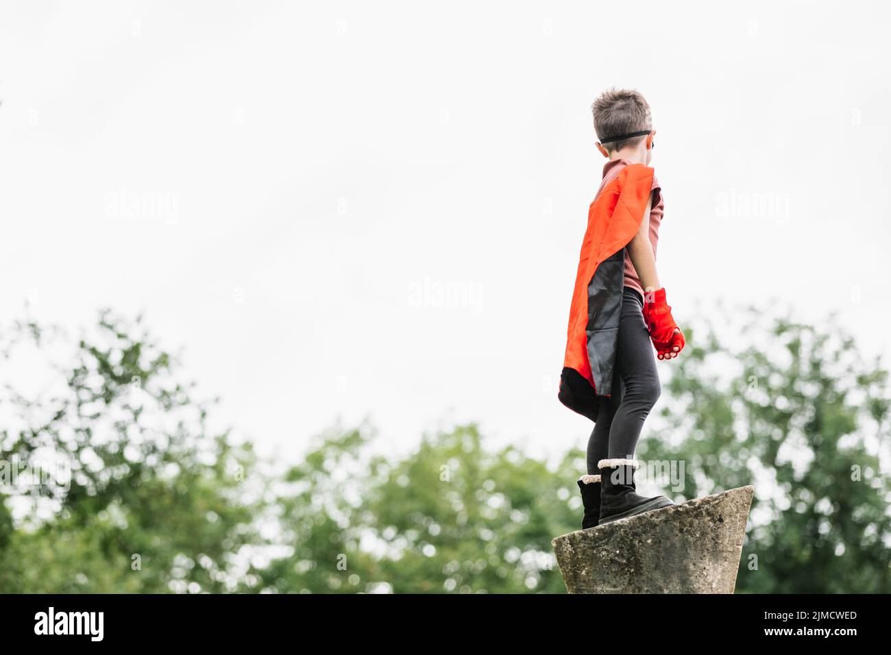 Side view of unrecognizable boy in red superhero cape and mask looking away while standing on stone block on blurred background of park trees Stock Photo