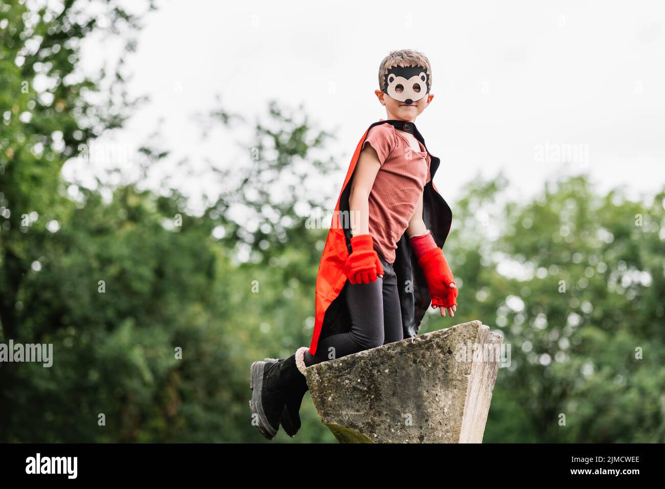 Side view of smiling boy in red superhero cape and hedgehog mask looking at camera while kneeling on stone block on blurred background of park trees Stock Photo