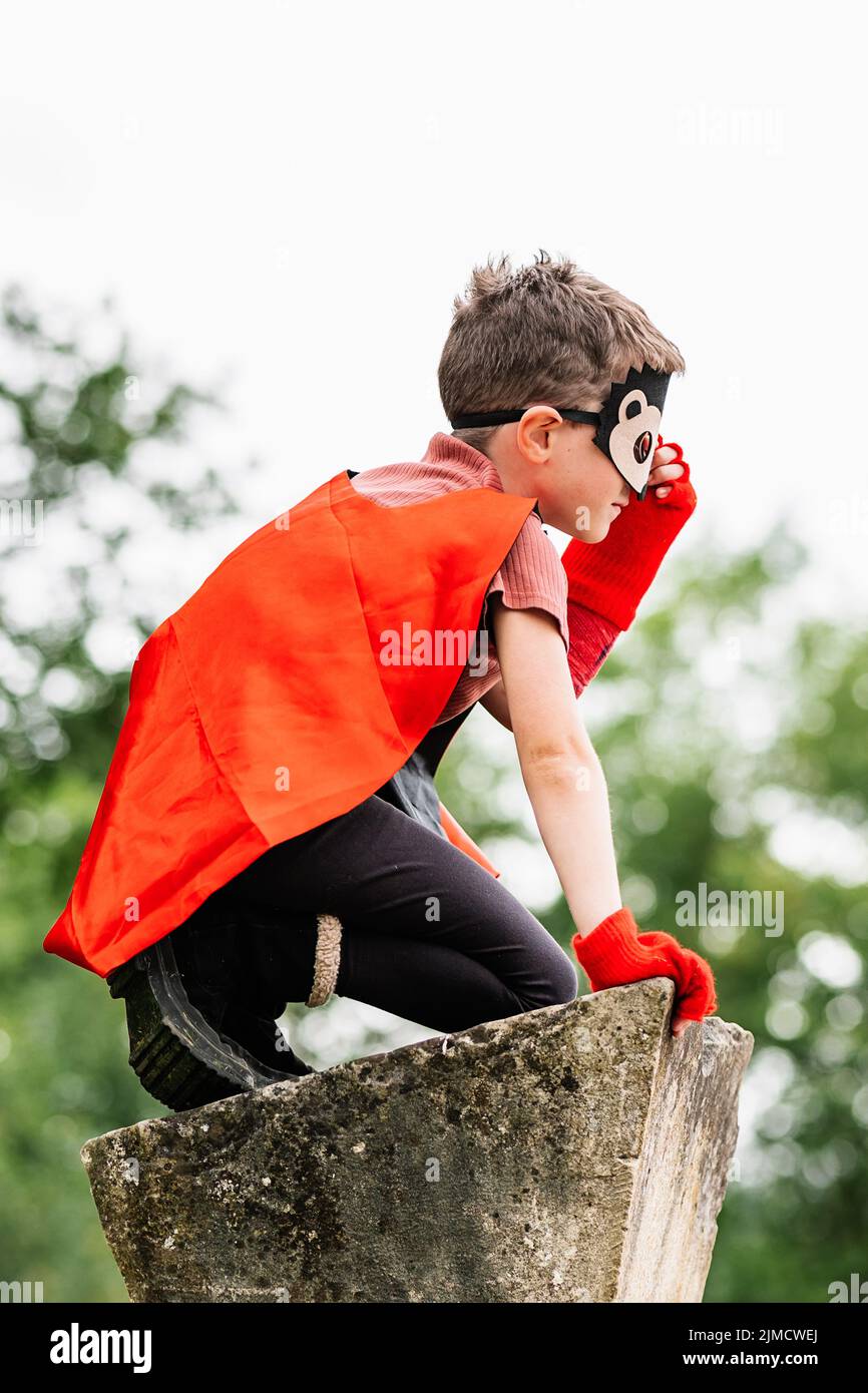 Side view of boy in red superhero cape and hedgehog mask touching face and looking away while kneeling on stone block on blurred background of park tr Stock Photo