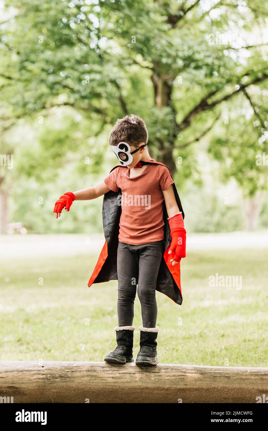 Full body cute little superhero boy with panda mask and cape walking on log while playing in green park in summer Stock Photo