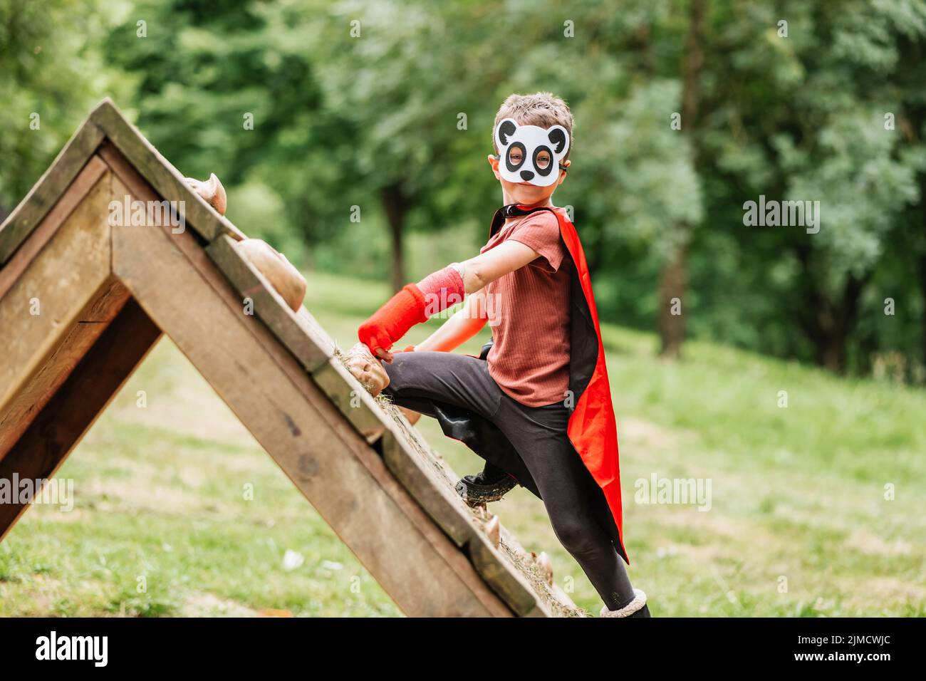 Side view of boy with red cape and panda mask climbing lumber wall looking at camera while pretending to be superhero on playground in park Stock Photo