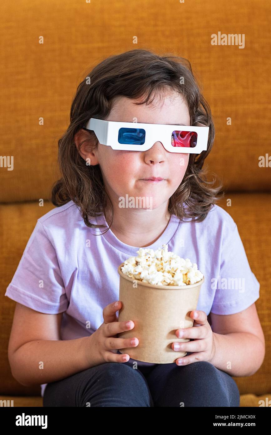 Kid watching movie in 3D glasses on couch with tasty popcorn in light