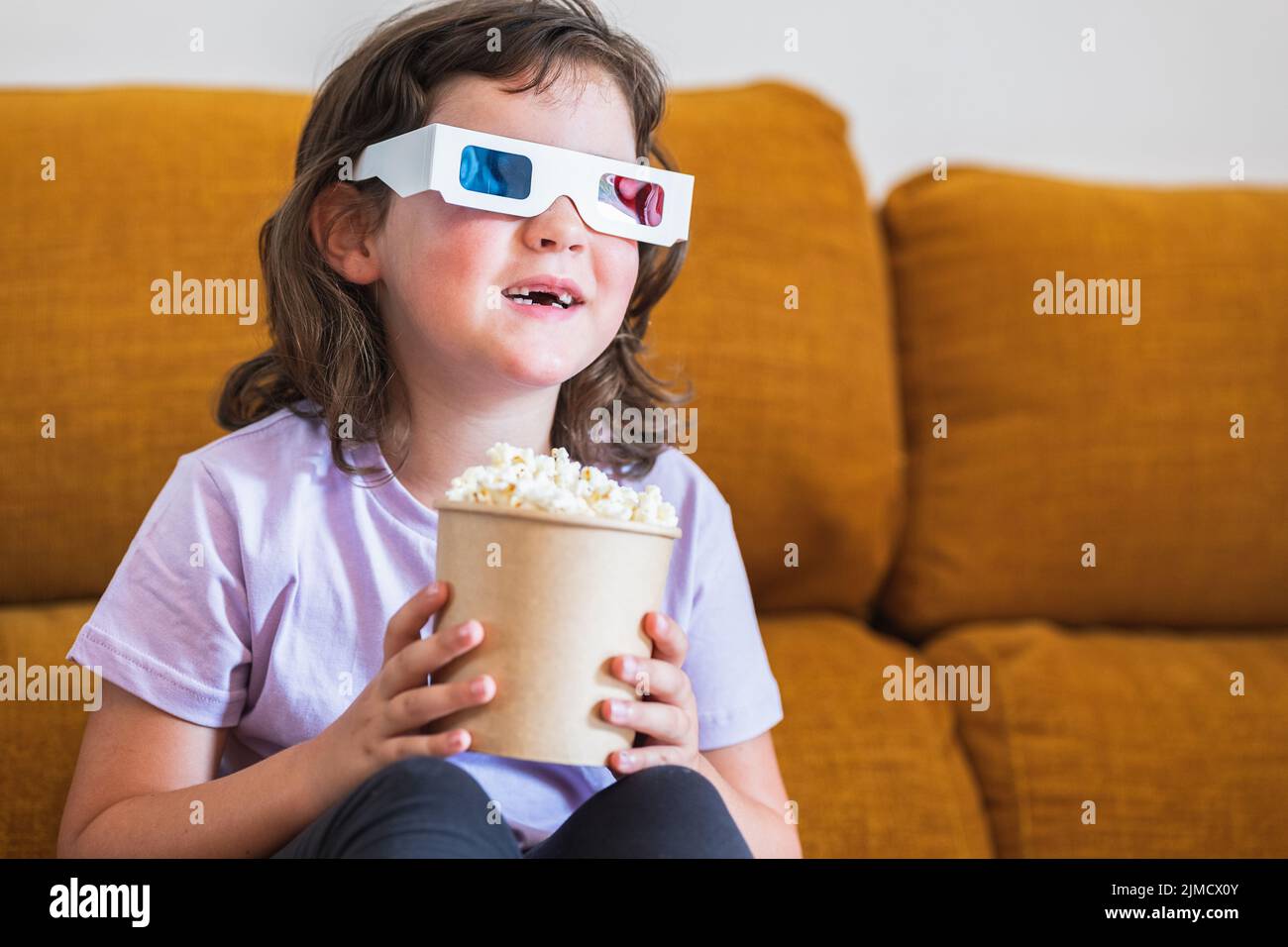 Cheerful kid watching movie in 3D glasses on couch with tasty popcorn in light living room at home Stock Photo