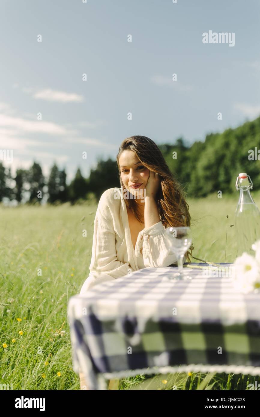 Female in dress looking at camera while sitting at table with beverage in grassy field on sunny summer day in countryside Stock Photo