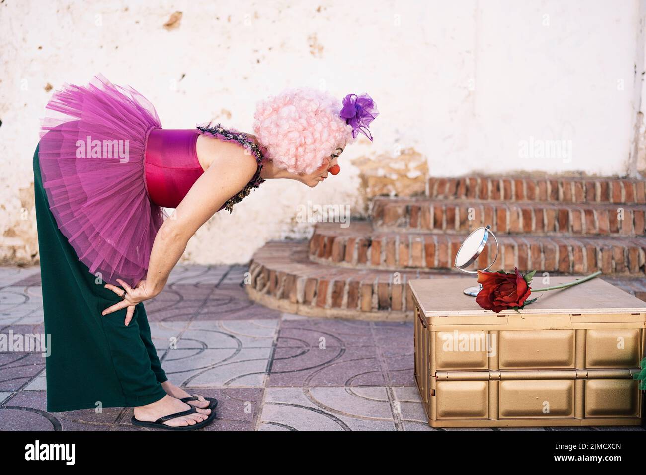 Side view of adult female artist in funny clown dress pink wig and nose leaning forward and looking in small round mirror before street performance Stock Photo