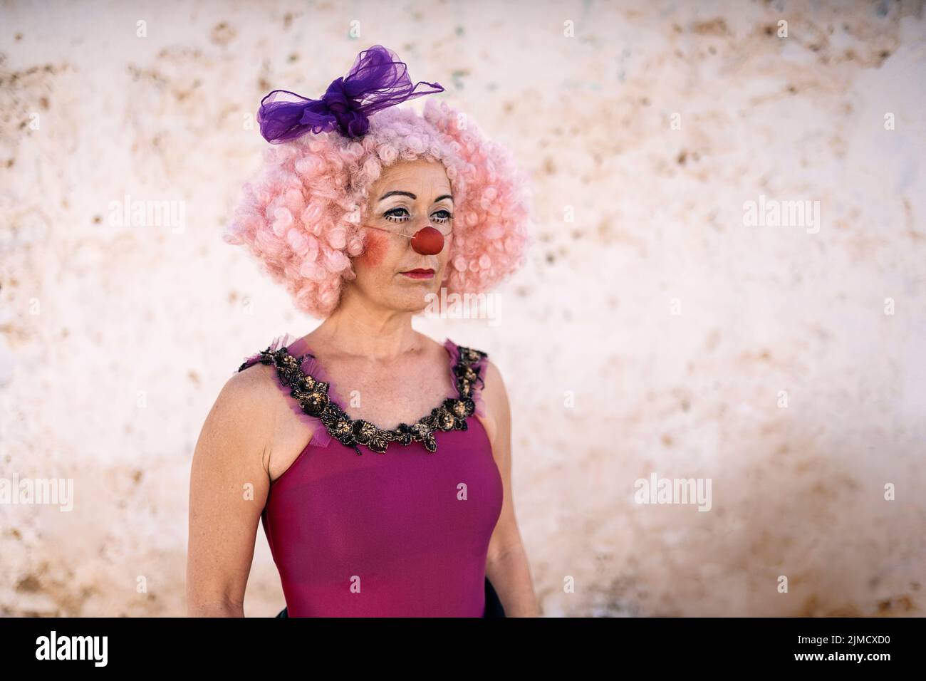 Concentrated middle aged female circus artist in funny costume and pink wig with bright makeup and fake nose standing near wall before performance on Stock Photo