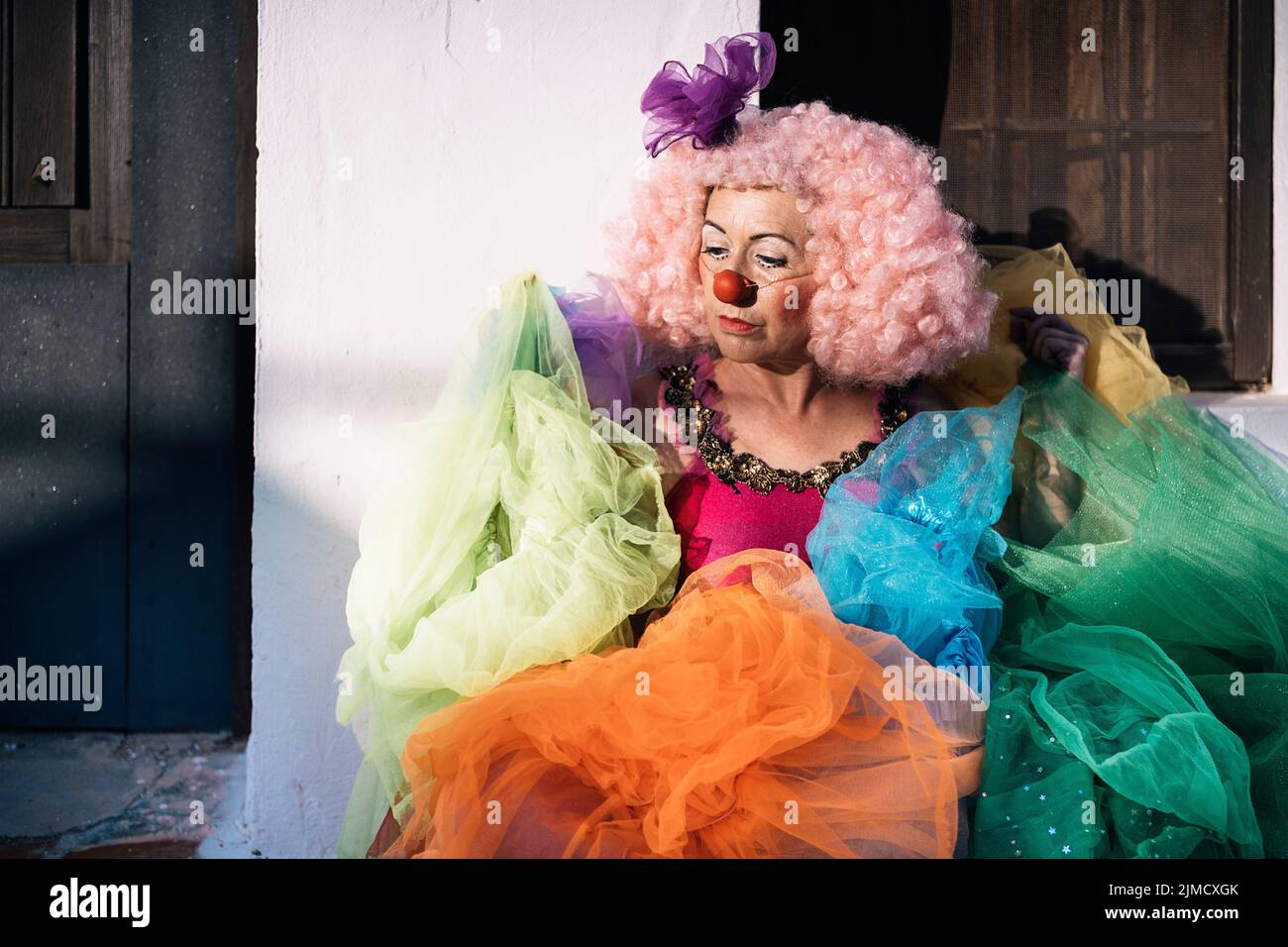 Side view of sad mature female clown in curly pink wig with bright makeup carrying pile of tutu skirts and looking away after performance on street on Stock Photo