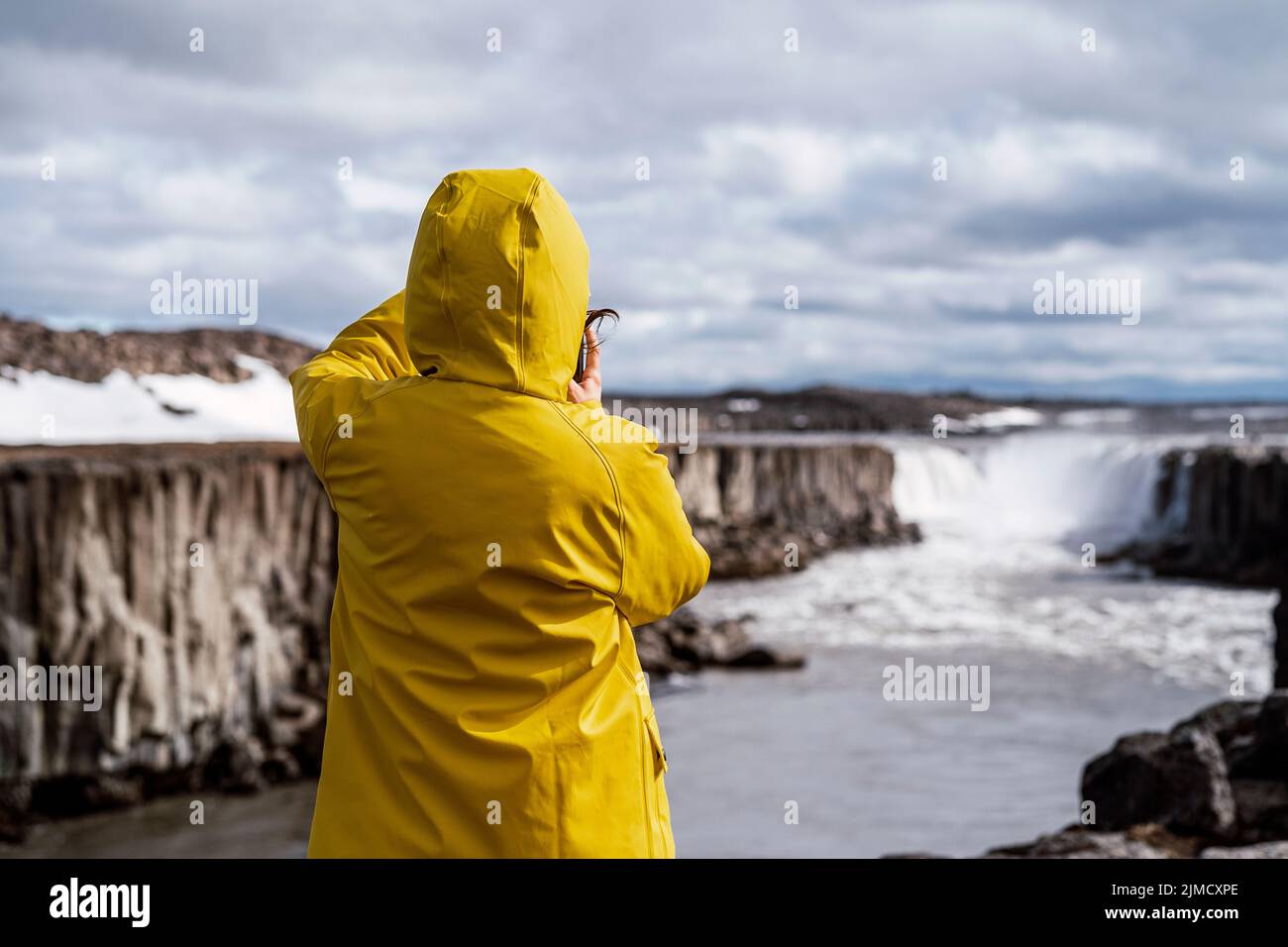 Back view of unrecognizable traveler in bright yellow hooded raincoat taking photo of amazing scenery of Selfoss waterfall and river flowing in gorge Stock Photo