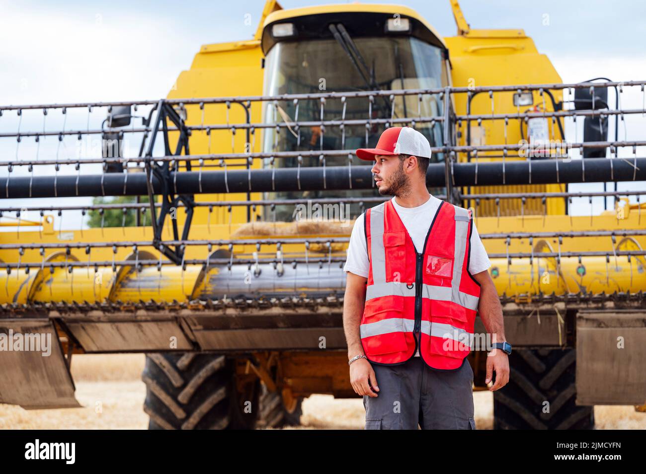 Bearded male farmer in uniform looking away while standing on agricultural field with industrial combine harvester during work in countryside Stock Photo