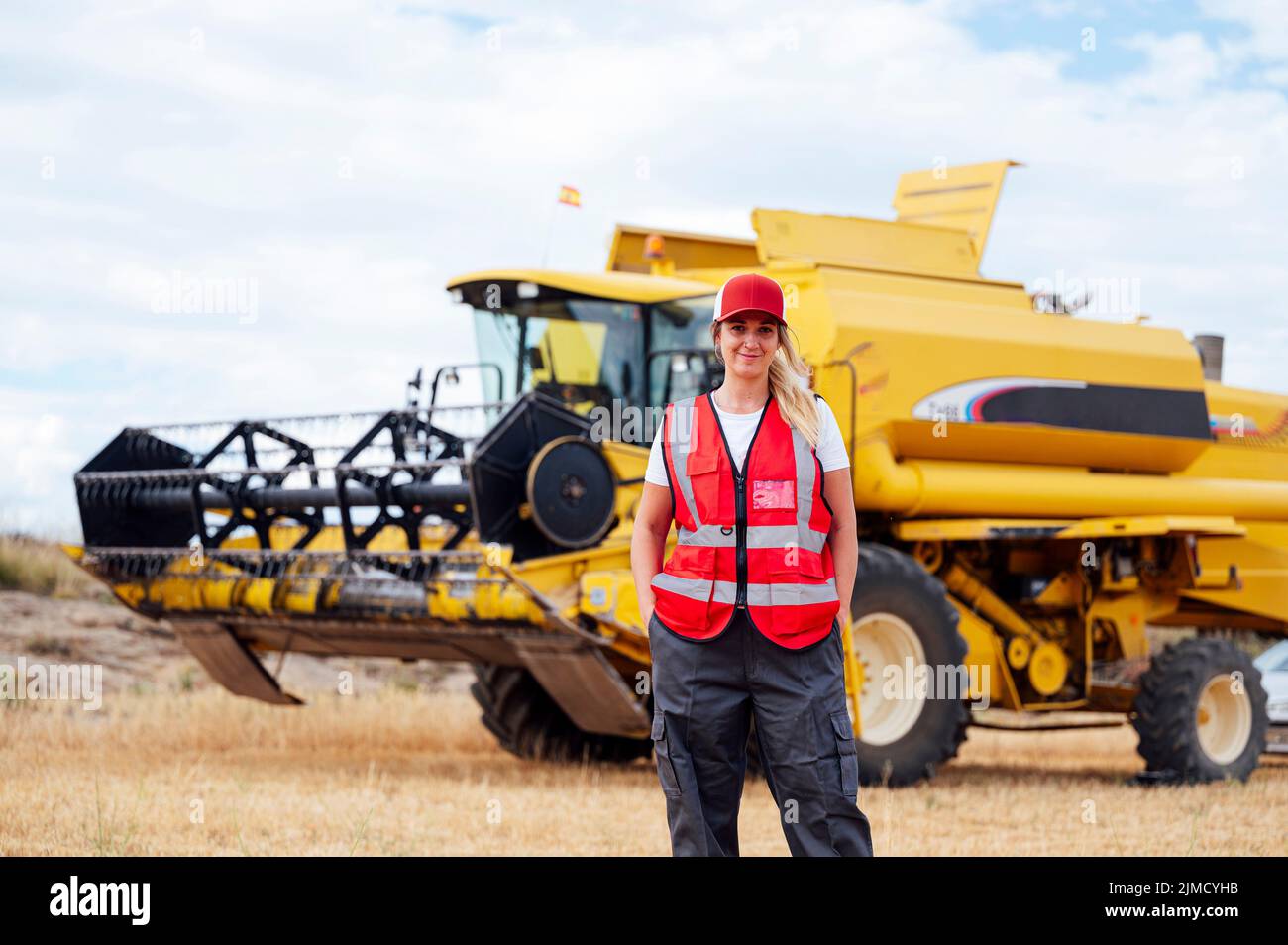Positive female worker in uniform looking at camera while standing in agricultural field with industrial combine harvester in rural area Stock Photo