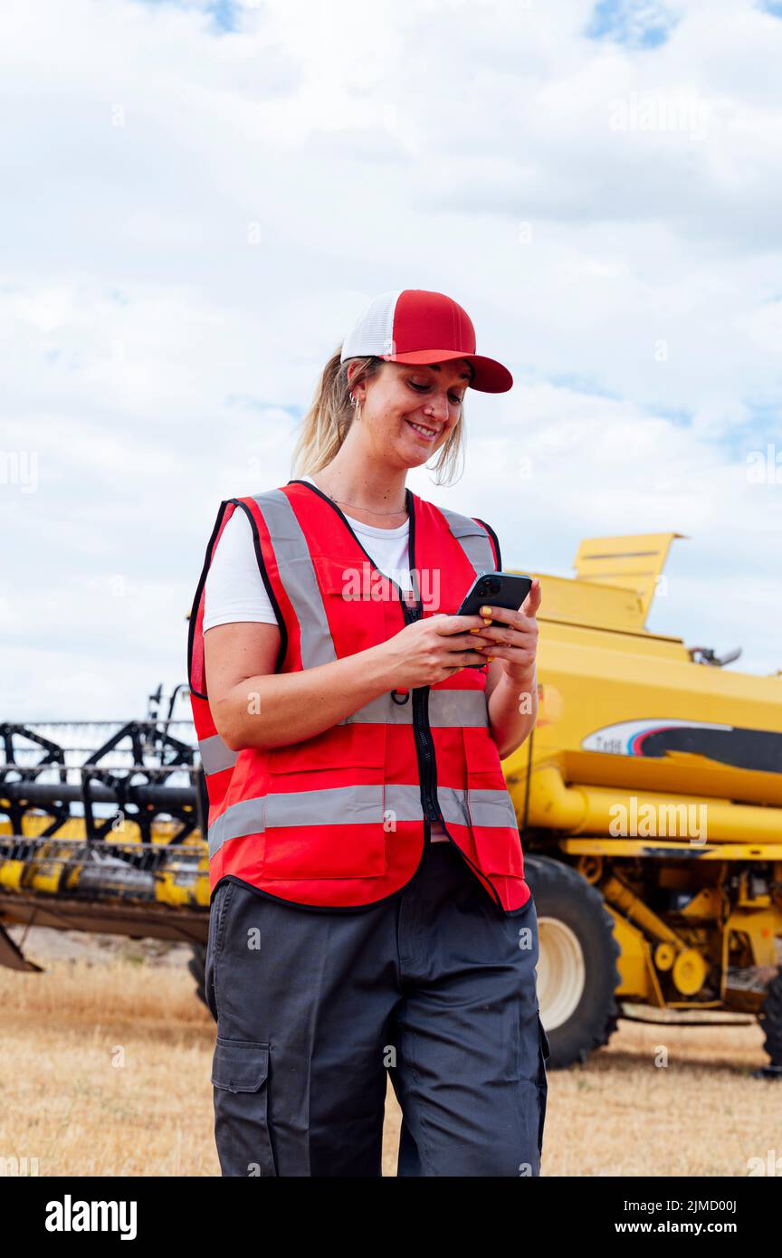 Cheerful female worker in uniform talking on cellphone while standing near industrial combine harvester during work in countryside on summer day Stock Photo