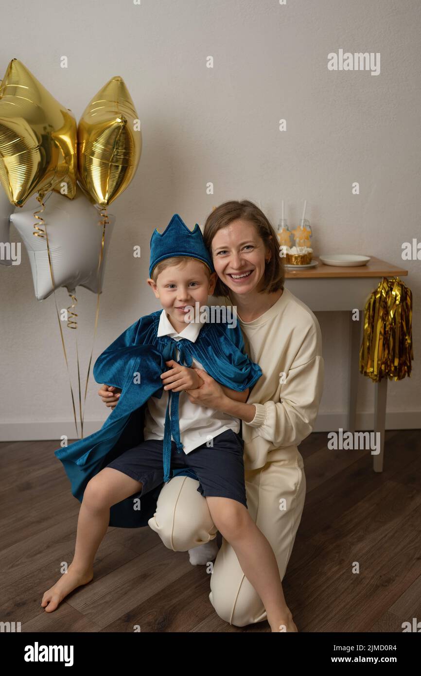 Smiling mother holding little boy dressed up in a blue cape and crown looking at camera at a birthday party at home Stock Photo