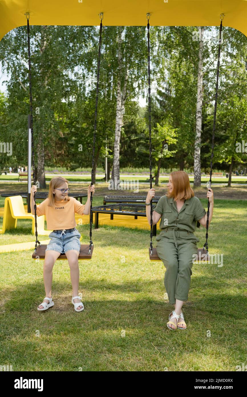Content girl looking at positive mother sitting on swing while spending time together in park with green trees on summer day Stock Photo